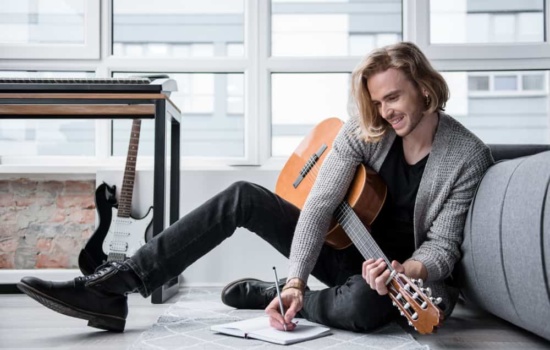 Young male musician sitting on floor with guitar and lyrics notebook